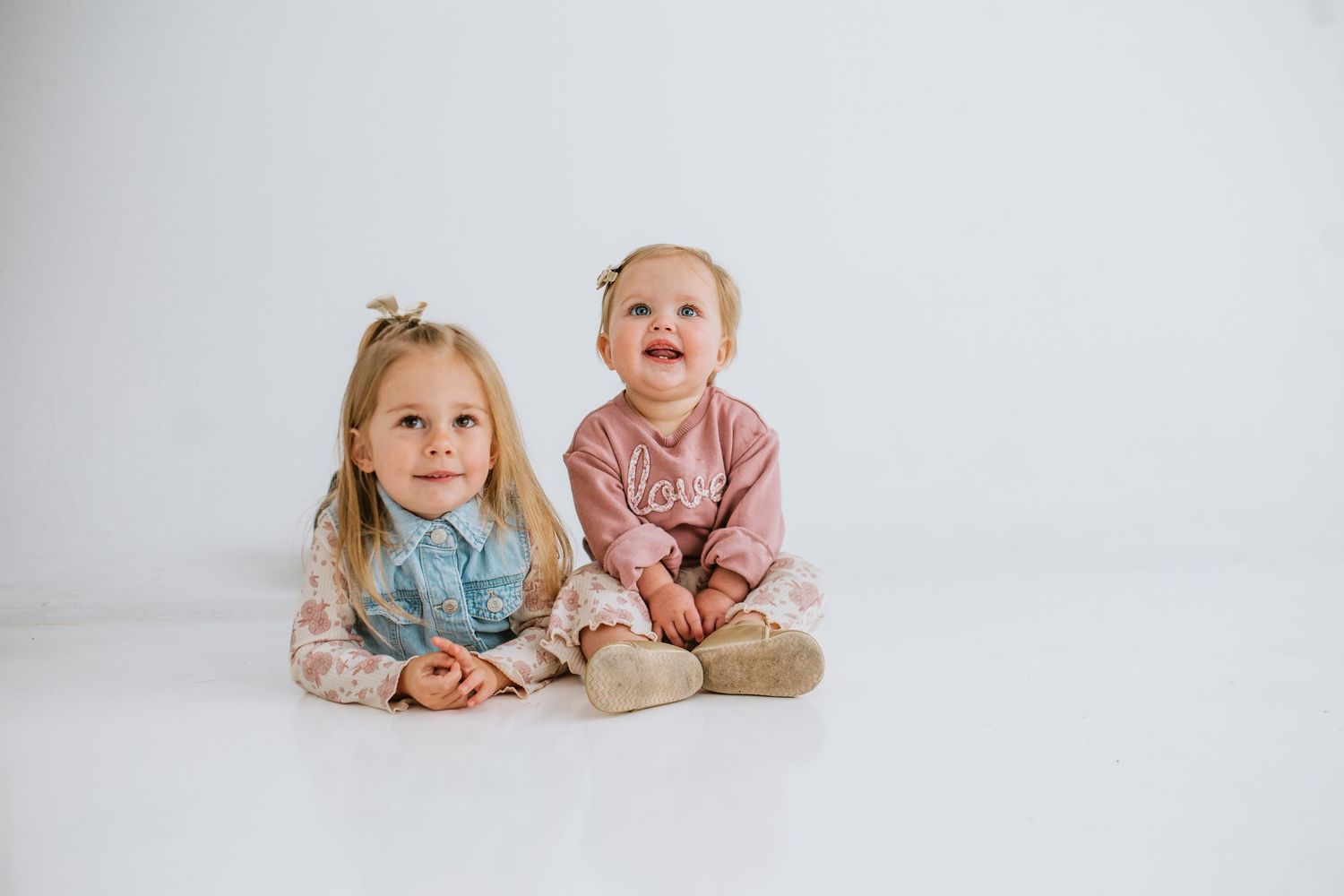 Two young sisters in pink outfits sitting together on a white floor, smiling cutely.