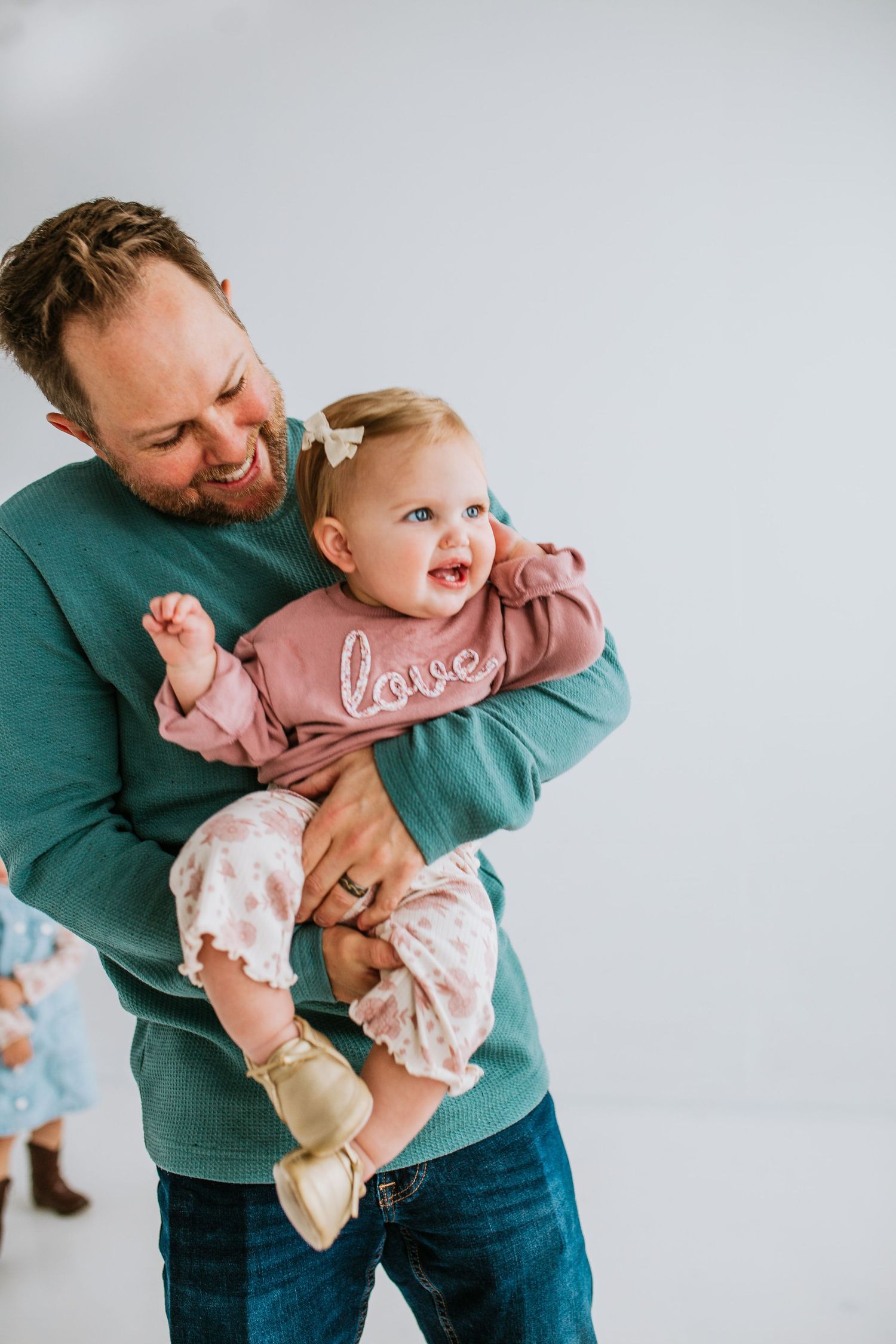 A bearded father in a teal shirt laughing while holding his baby girl in a pink love outfit.