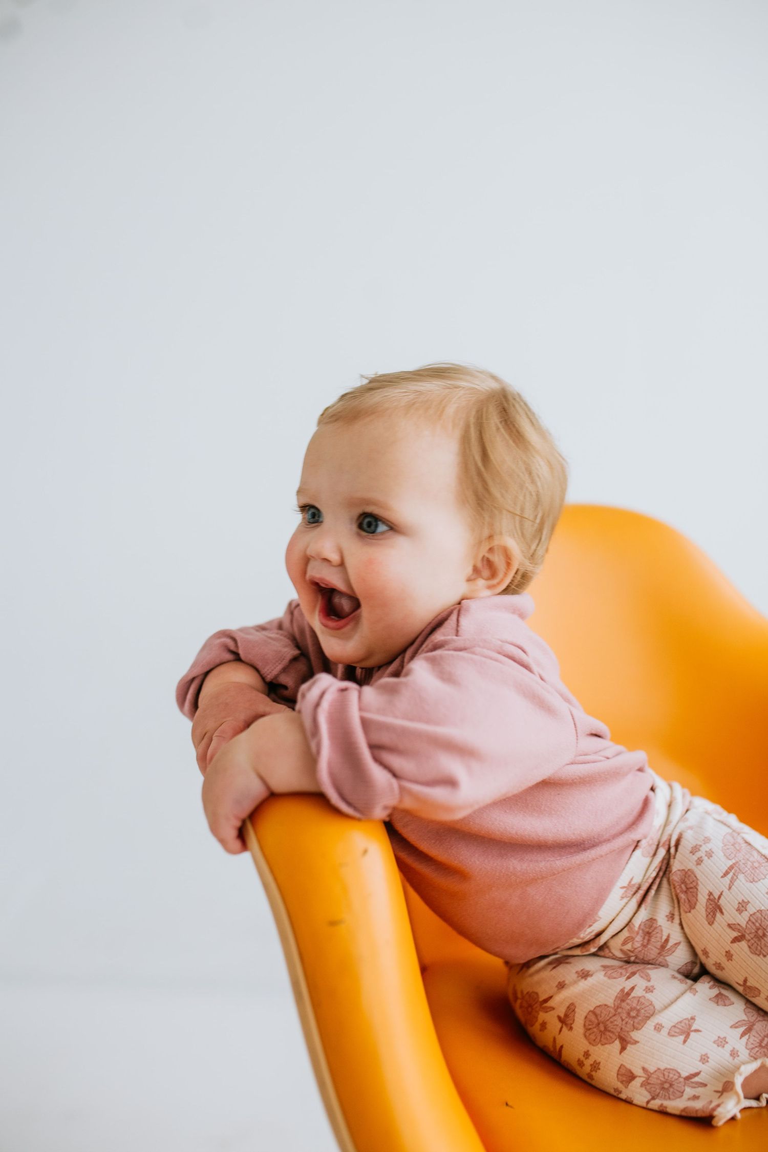 Happy baby in pink outfit sitting on a bright orange chair, laughing with arms resting on the chair back.