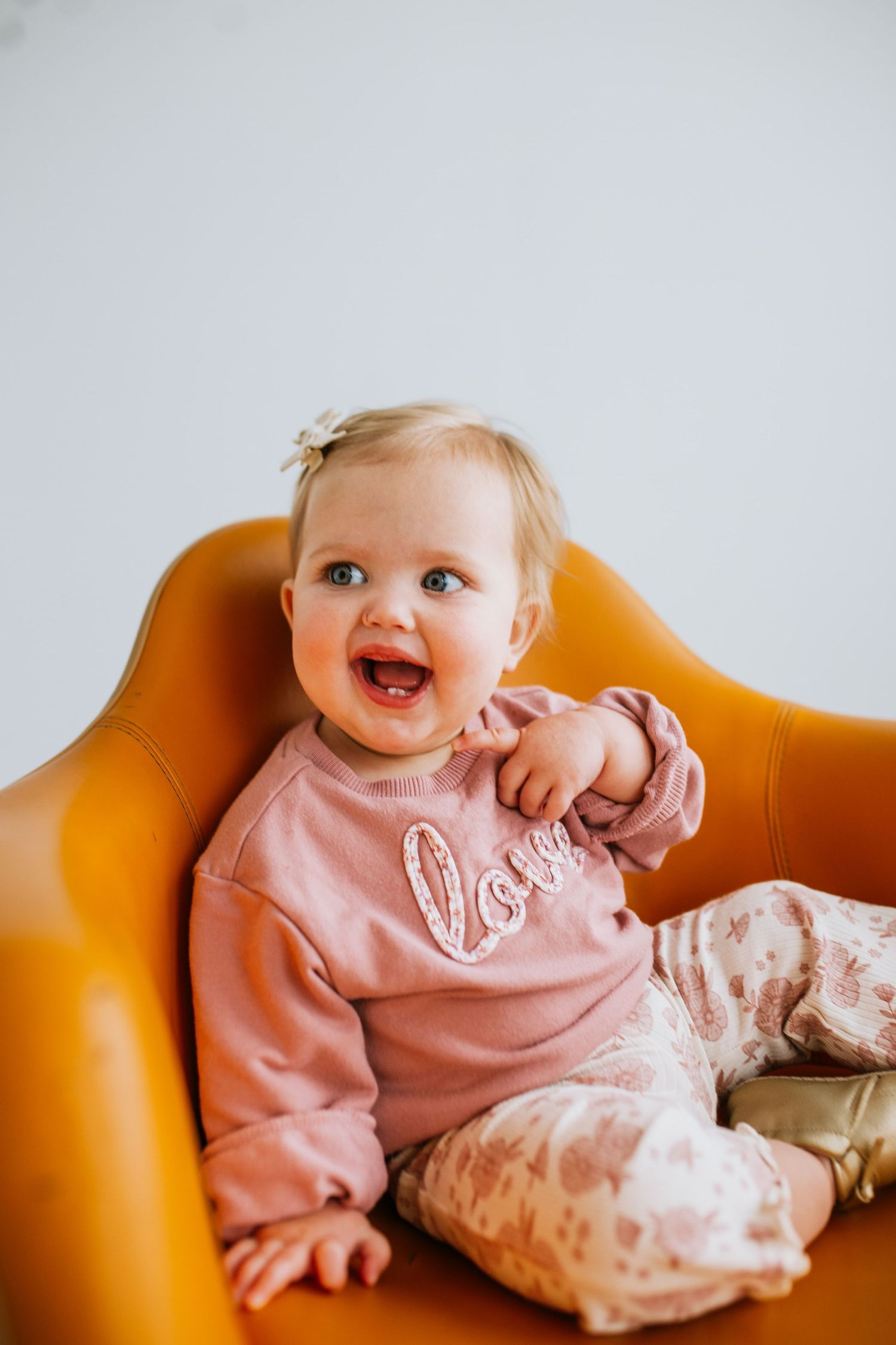 Adorable baby girl in pink love sweatshirt sitting in orange chair, smiling happily with blue eyes and hair bow.