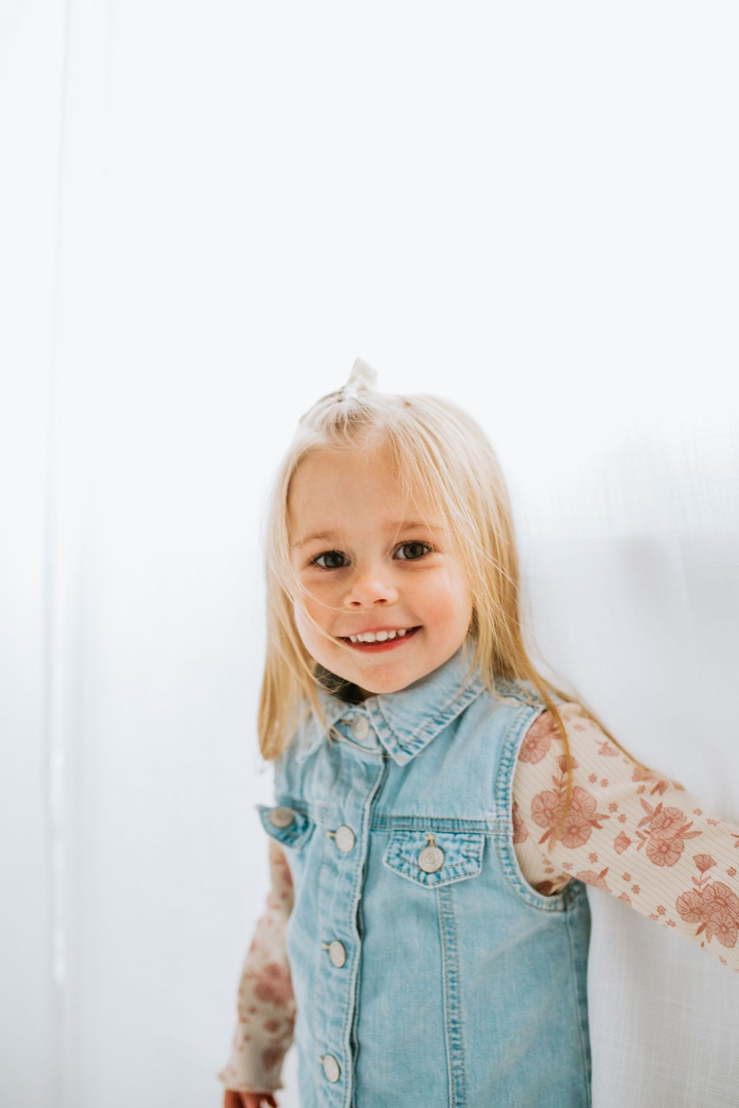 Smiling blonde toddler girl in denim vest and floral shirt with hair bow poses against white background.