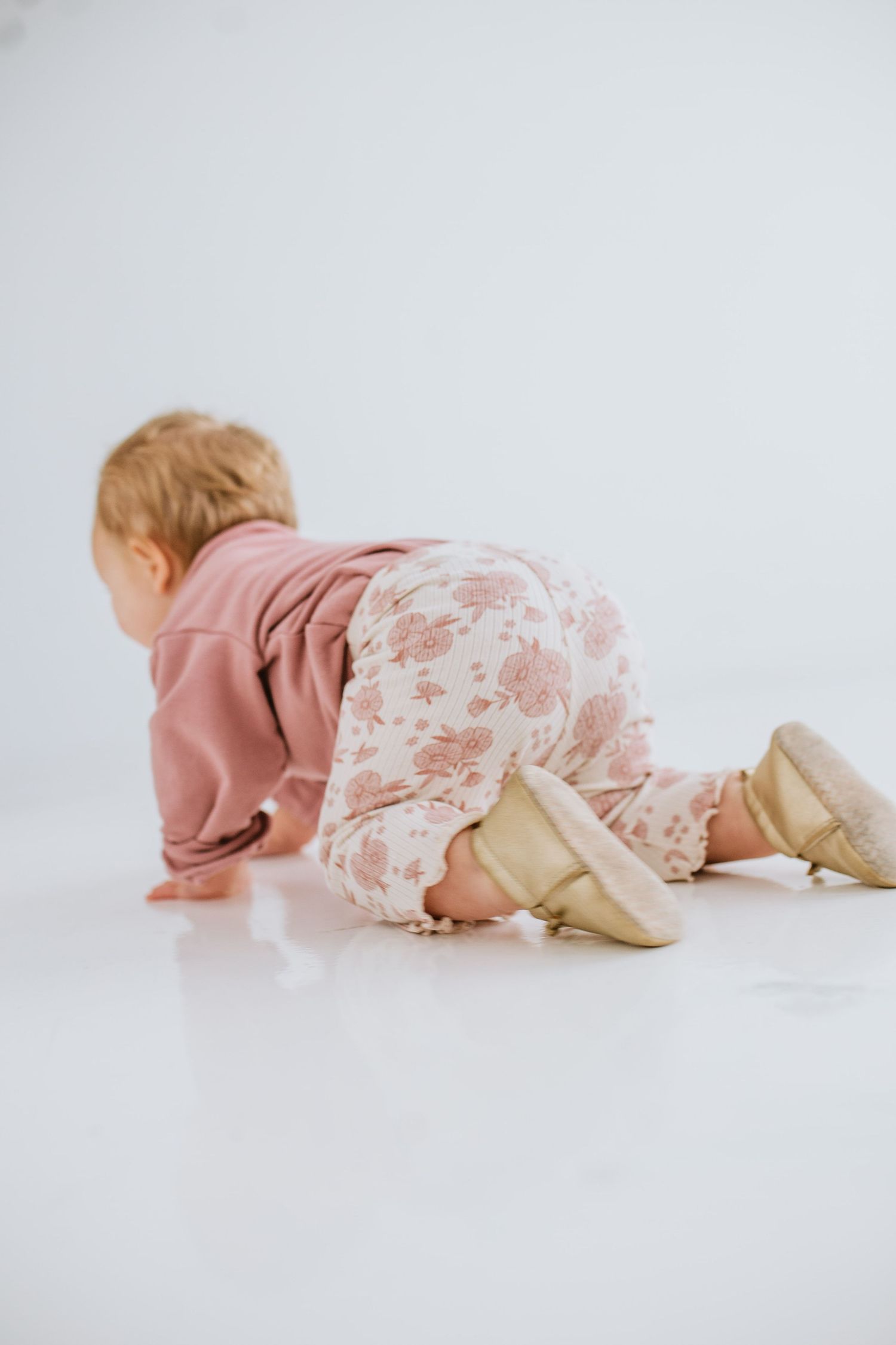 A baby in a floral outfit and gold booties crawling on a white floor.