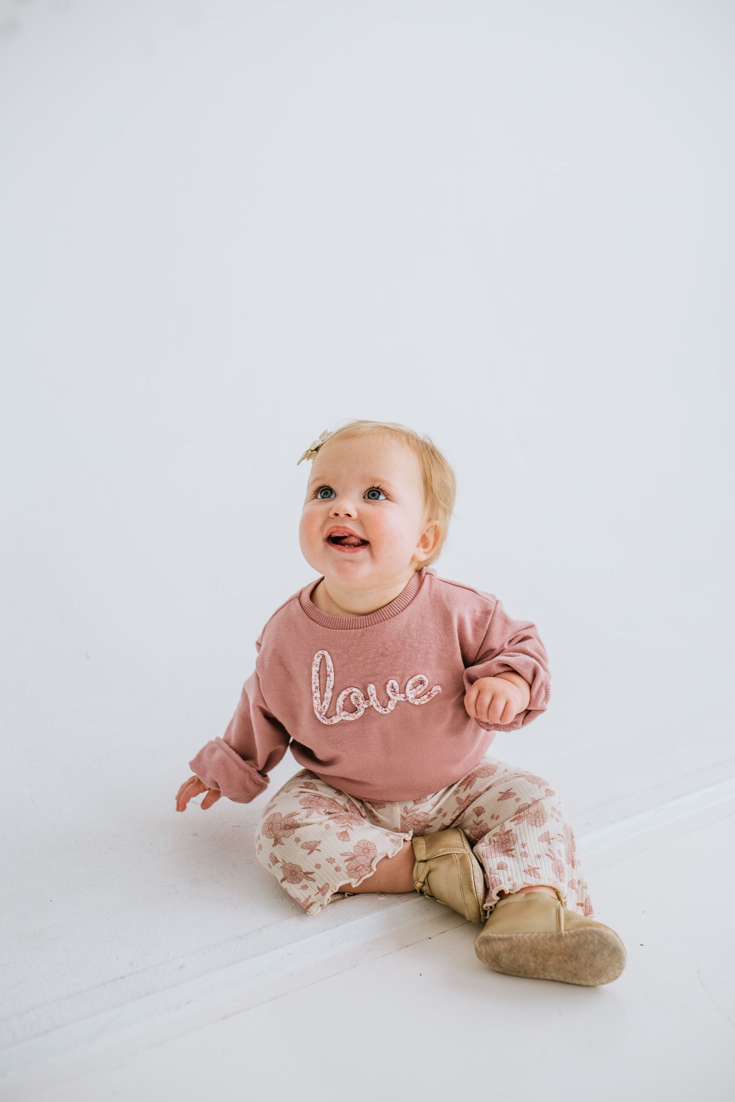 Adorable baby girl in a pink love sweatshirt and floral pants sitting on a white floor smiling.