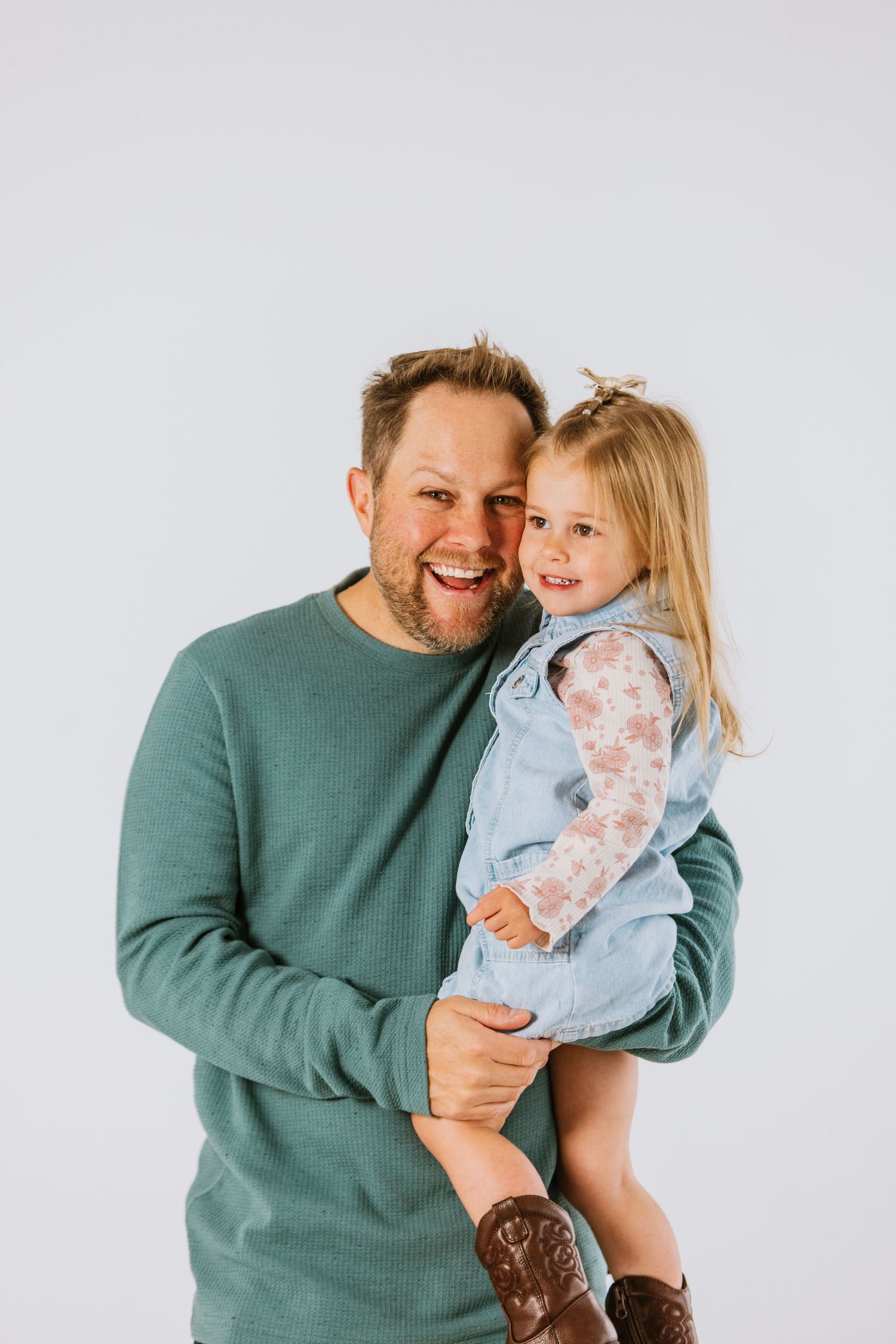 Happy father in teal sweater holds laughing blonde toddler daughter in denim vest against white backdrop.