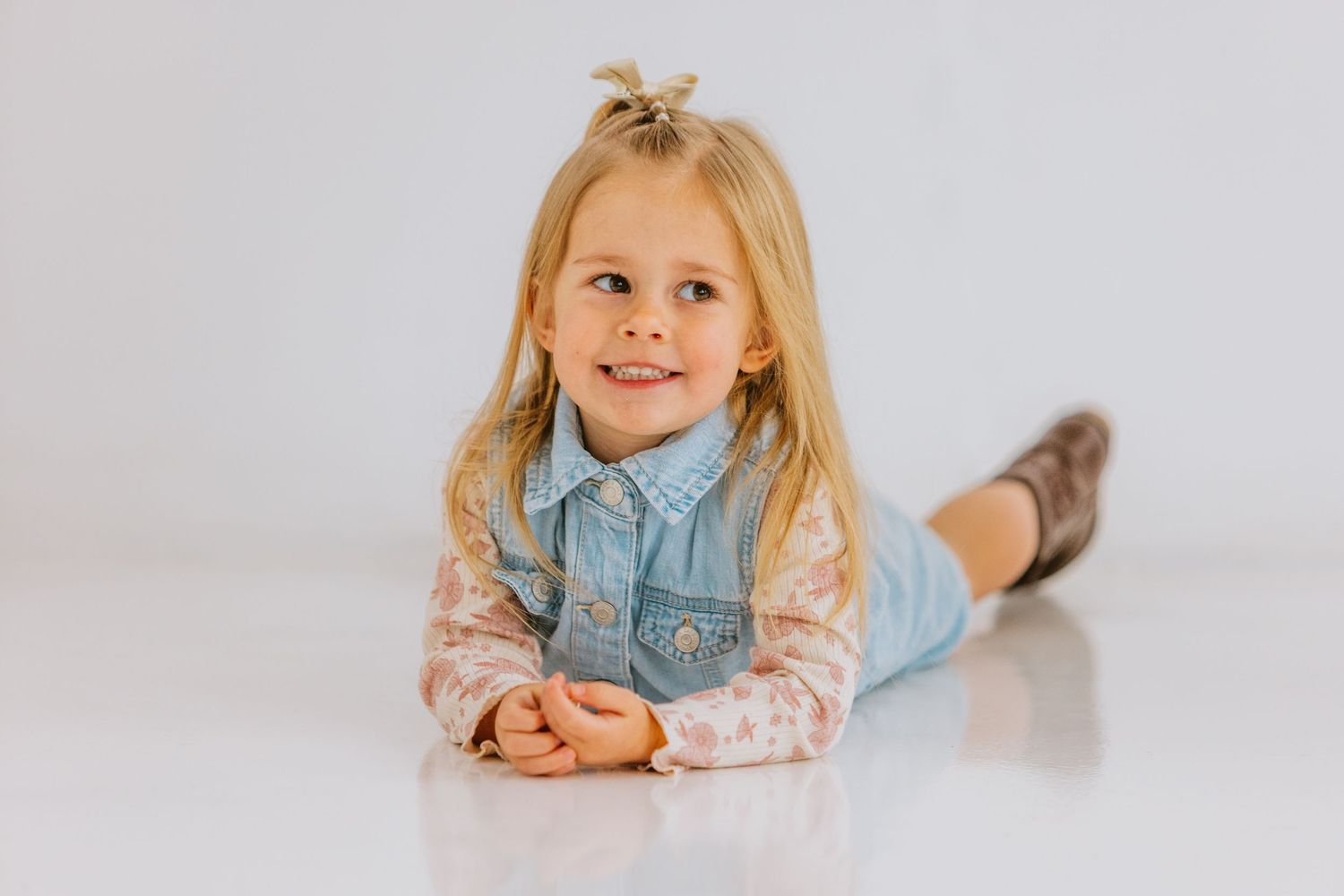 A young blonde girl in a denim vest lying on a white floor smiling.