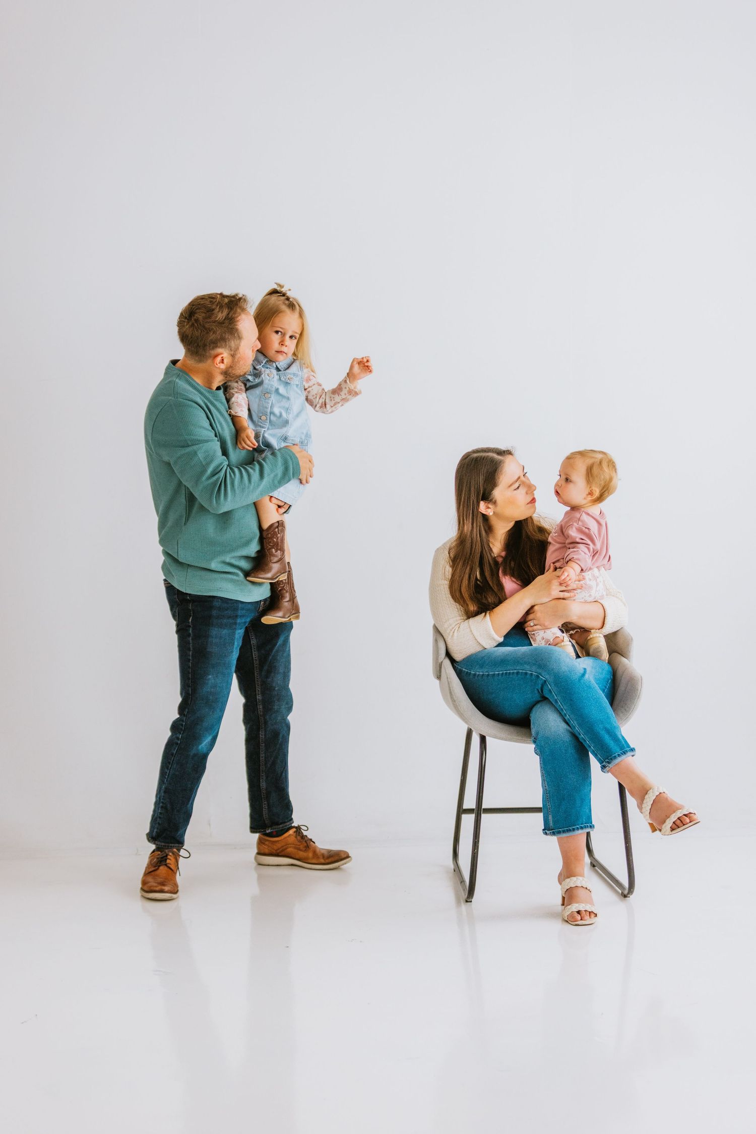 A young family with two toddlers poses in a bright white studio, parents each holding a child while smiling warmly.