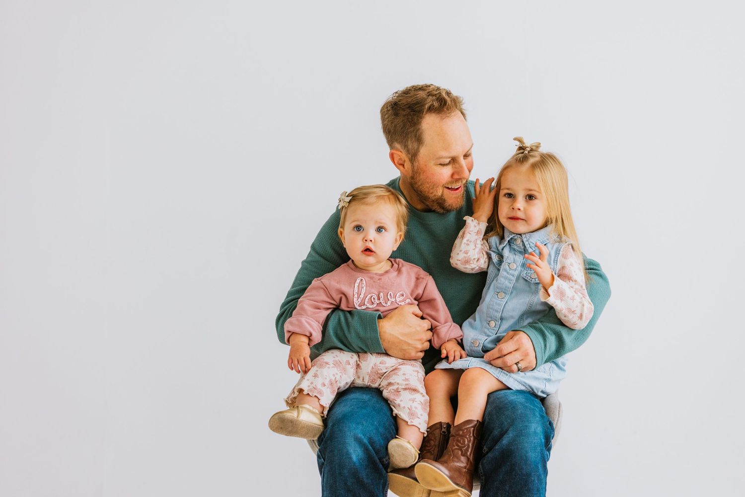 A red-haired father sits holding two young toddler girls in a bright white studio during a family photo session.