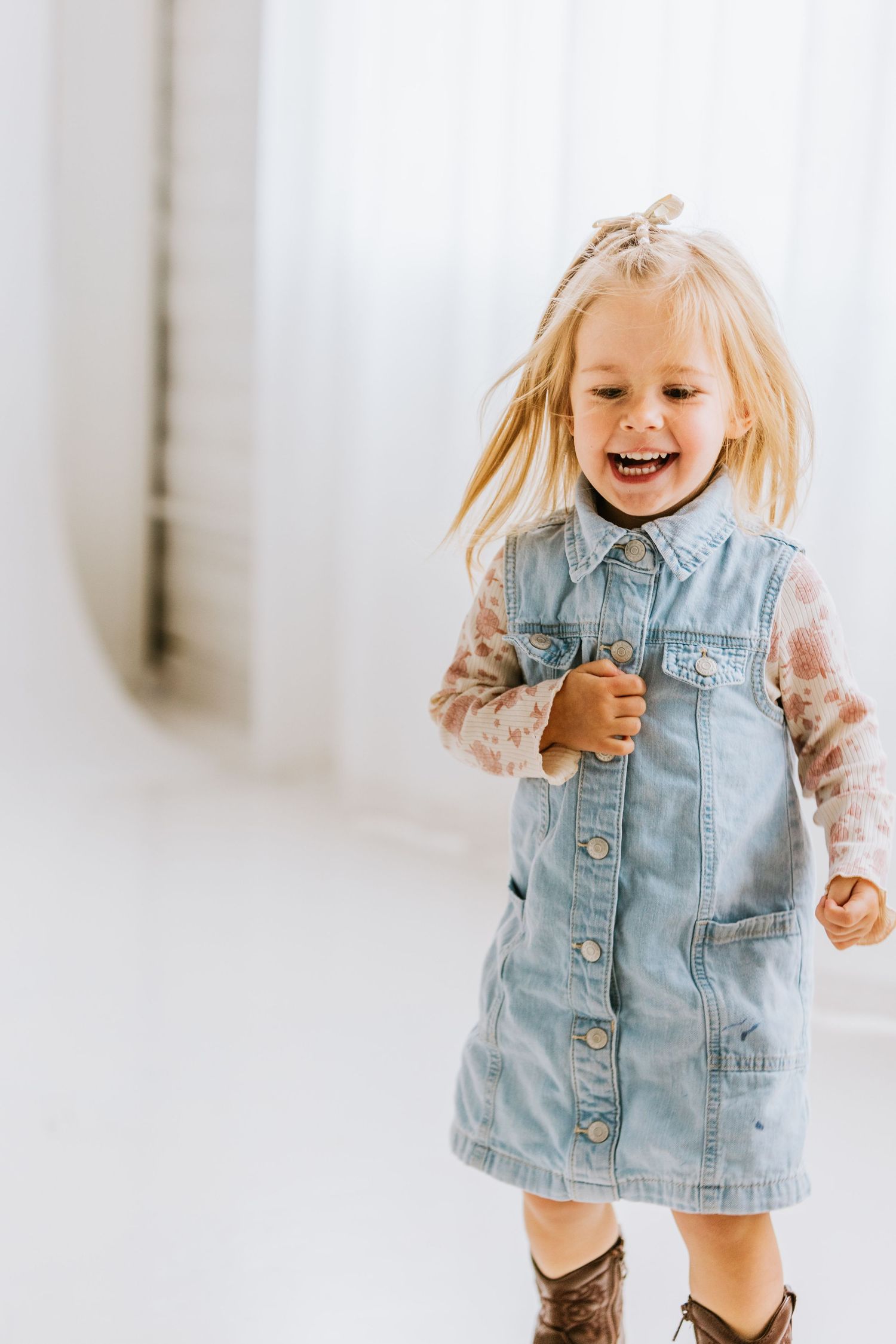 A joyful toddler girl in a denim dress and floral top laughs while playing indoors.