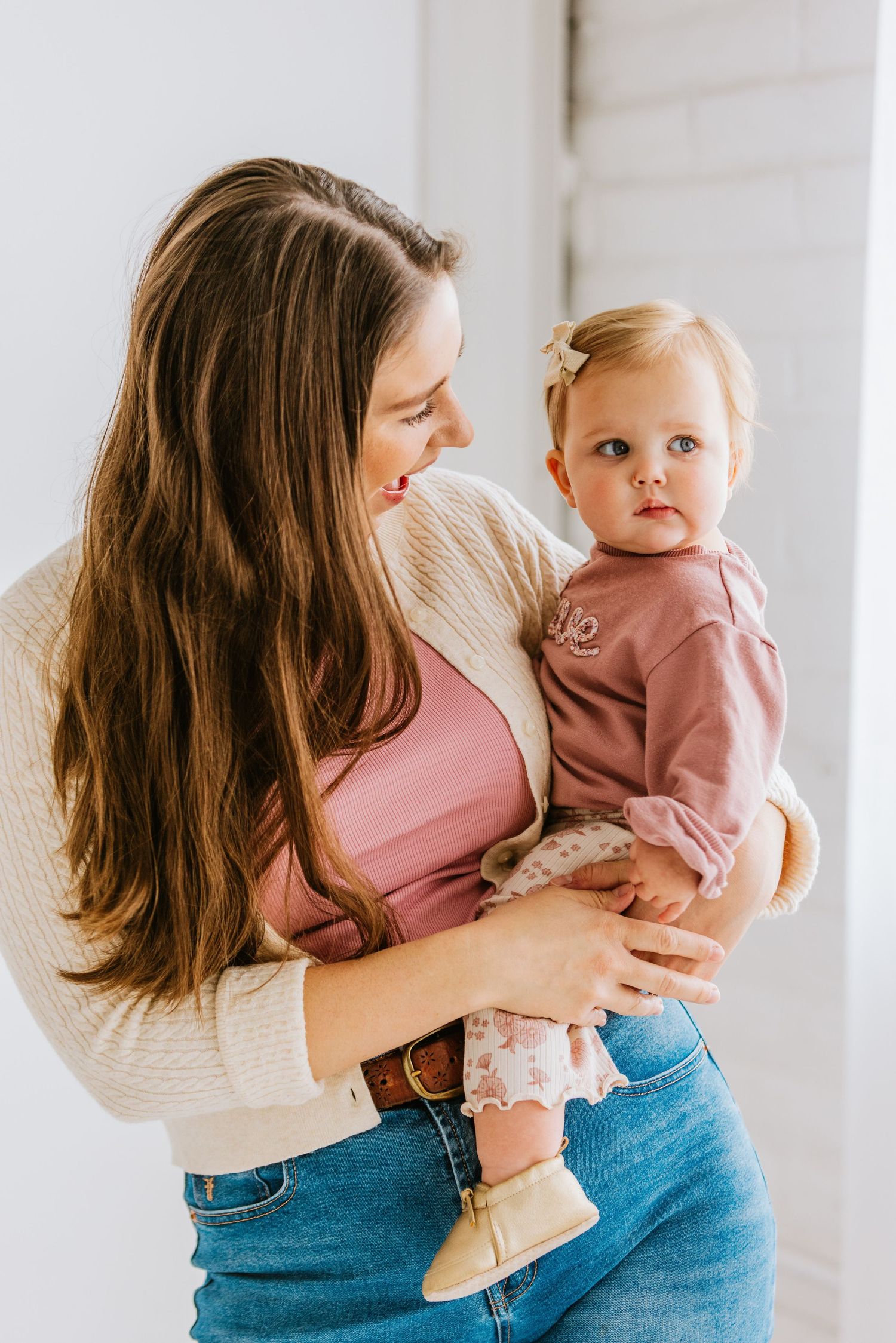 A young mother with long brown hair holds her baby girl in a bright white studio setting.
