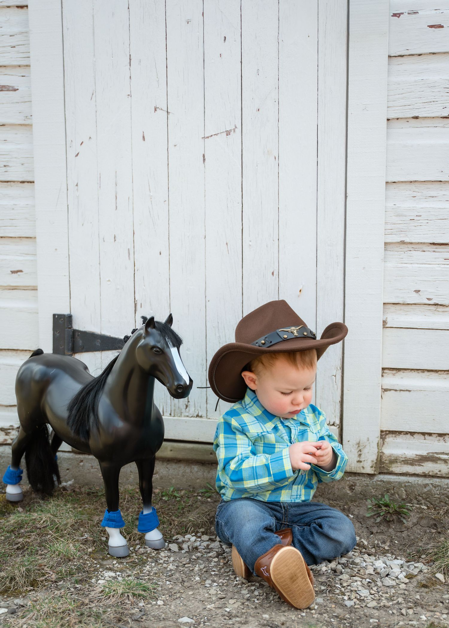 Toddler in cowboy hat and plaid shirt sits beside a black toy horse near a white barn door.