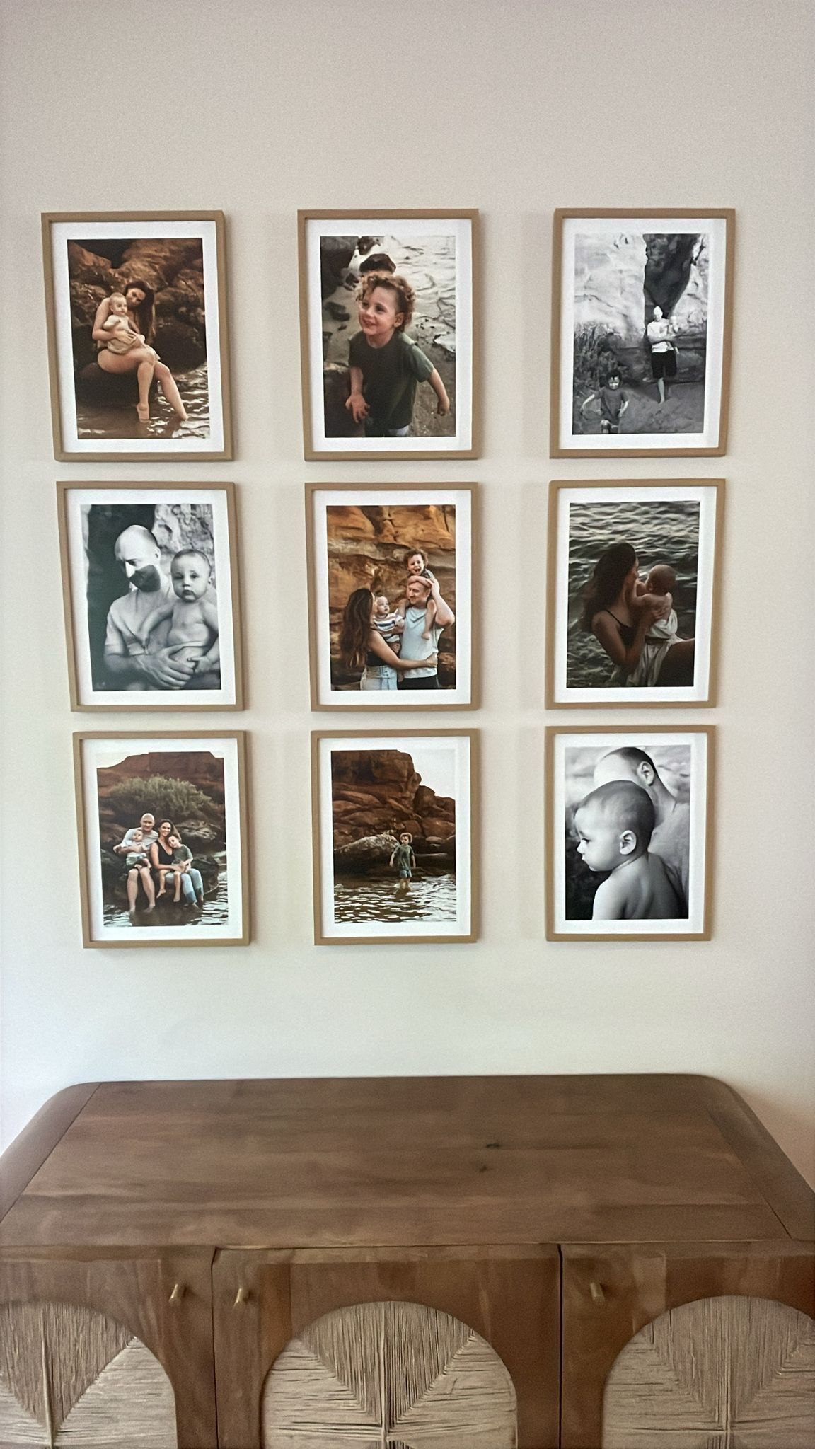 Nine framed family photos arranged in a 3x3 grid on a white wall above a wooden sideboard with wicker chairs.