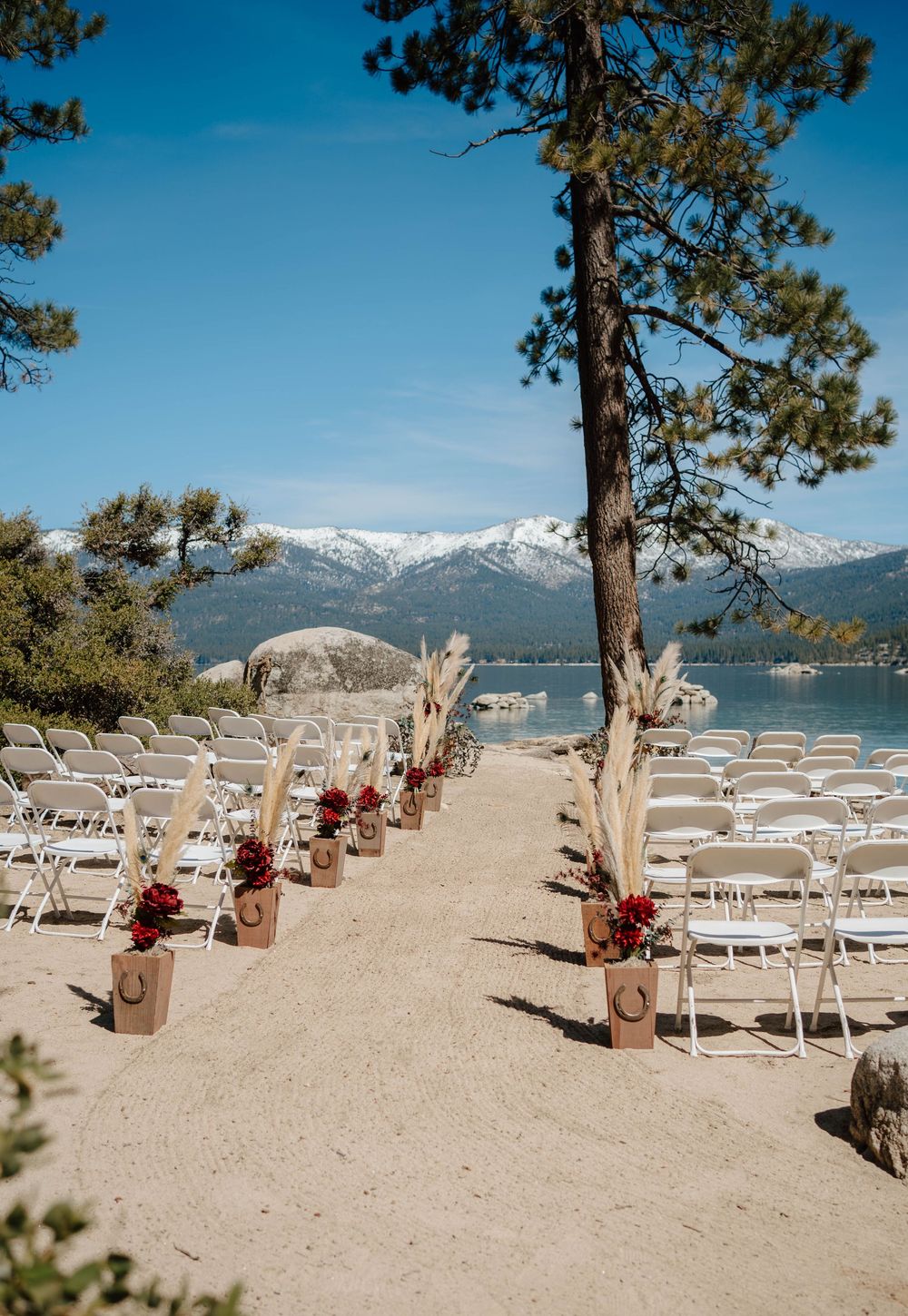 Outdoor lakeside wedding ceremony setup with white chairs, red floral arrangements, and snow-capped mountains in the background.