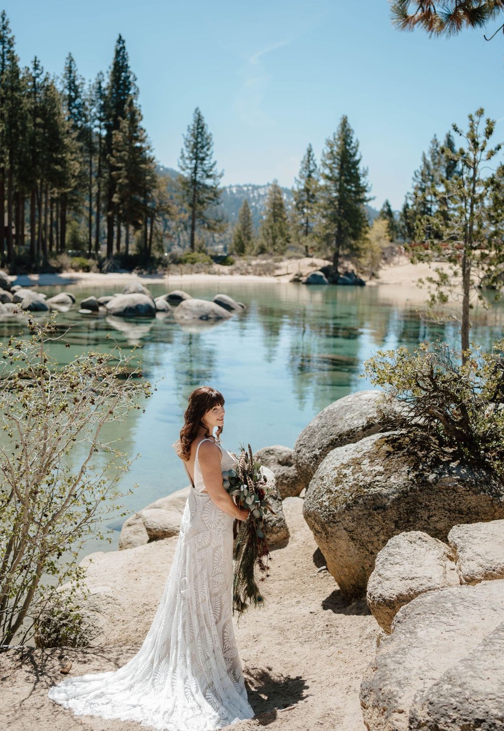 Bride in lace wedding gown holding bouquet stands among boulders beside a serene mountain lake surrounded by pine trees.