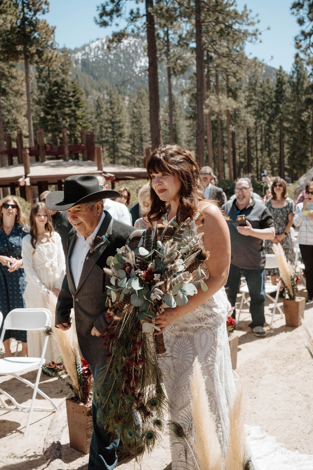 Bride in lace gown walks down outdoor aisle with older man in cowboy hat, holding wild floral bouquet, pine trees behind.