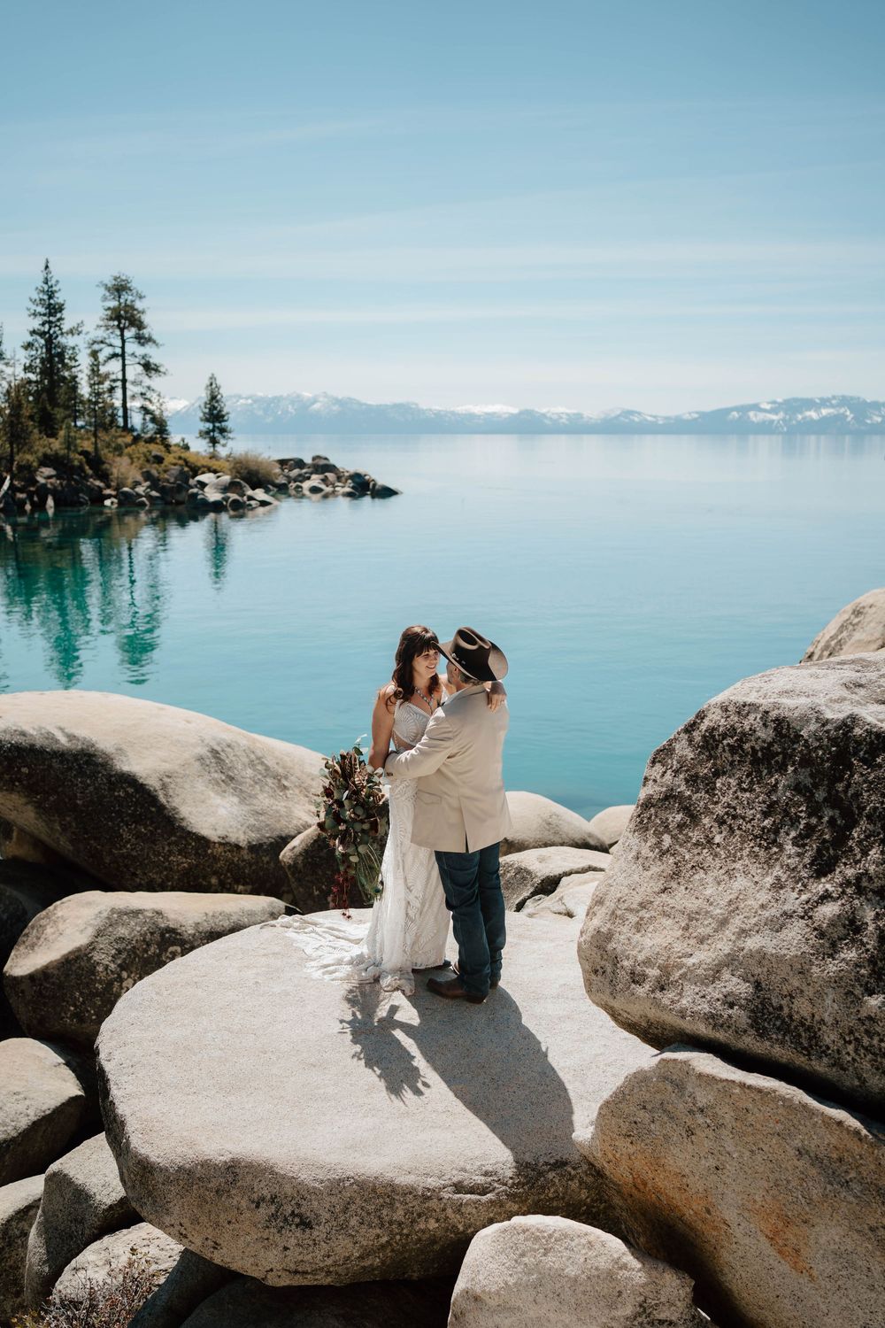 A couple shares a romantic embrace on lakeside boulders with turquoise water and mountains behind.