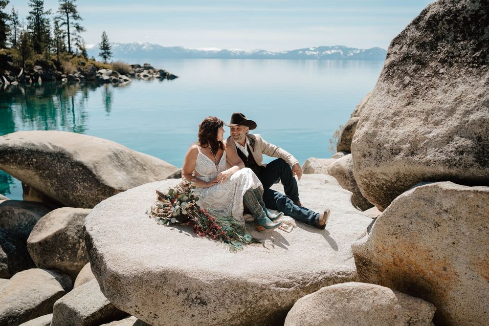 Couple in wedding attire sitting on large boulders at Lake Tahoe with turquoise water and mountains behind them.