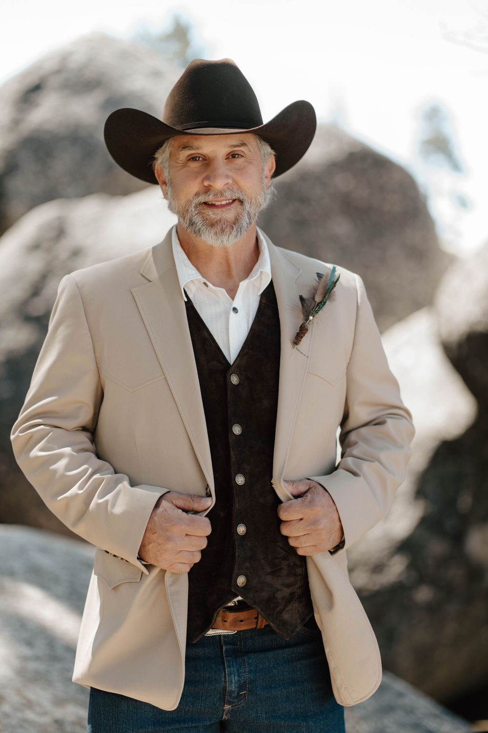 A groom in a black cowboy hat, tan blazer, and brown vest with a boutonniere poses on rocks.