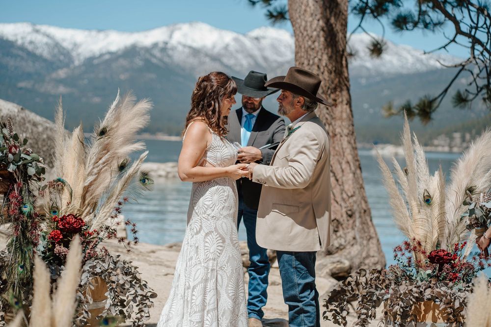 Couple exchanges vows at outdoor lakeside wedding ceremony with snowy mountains, pampas grass, and red floral arrangements.