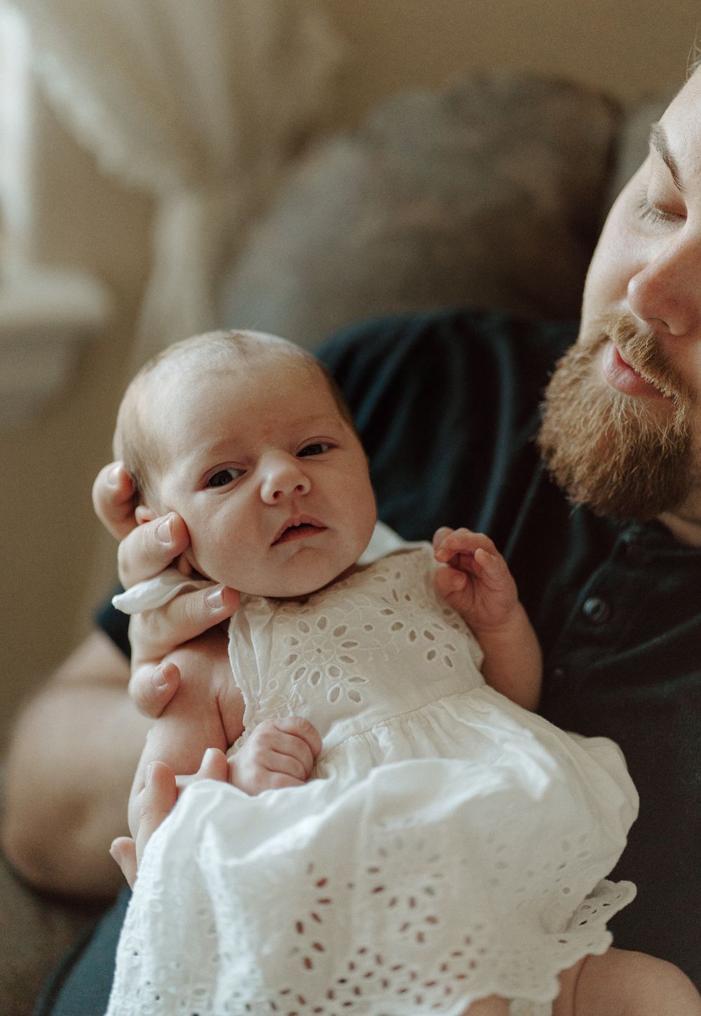 Bearded father tenderly holds newborn baby in white eyelet dress, sharing a gentle moment together.