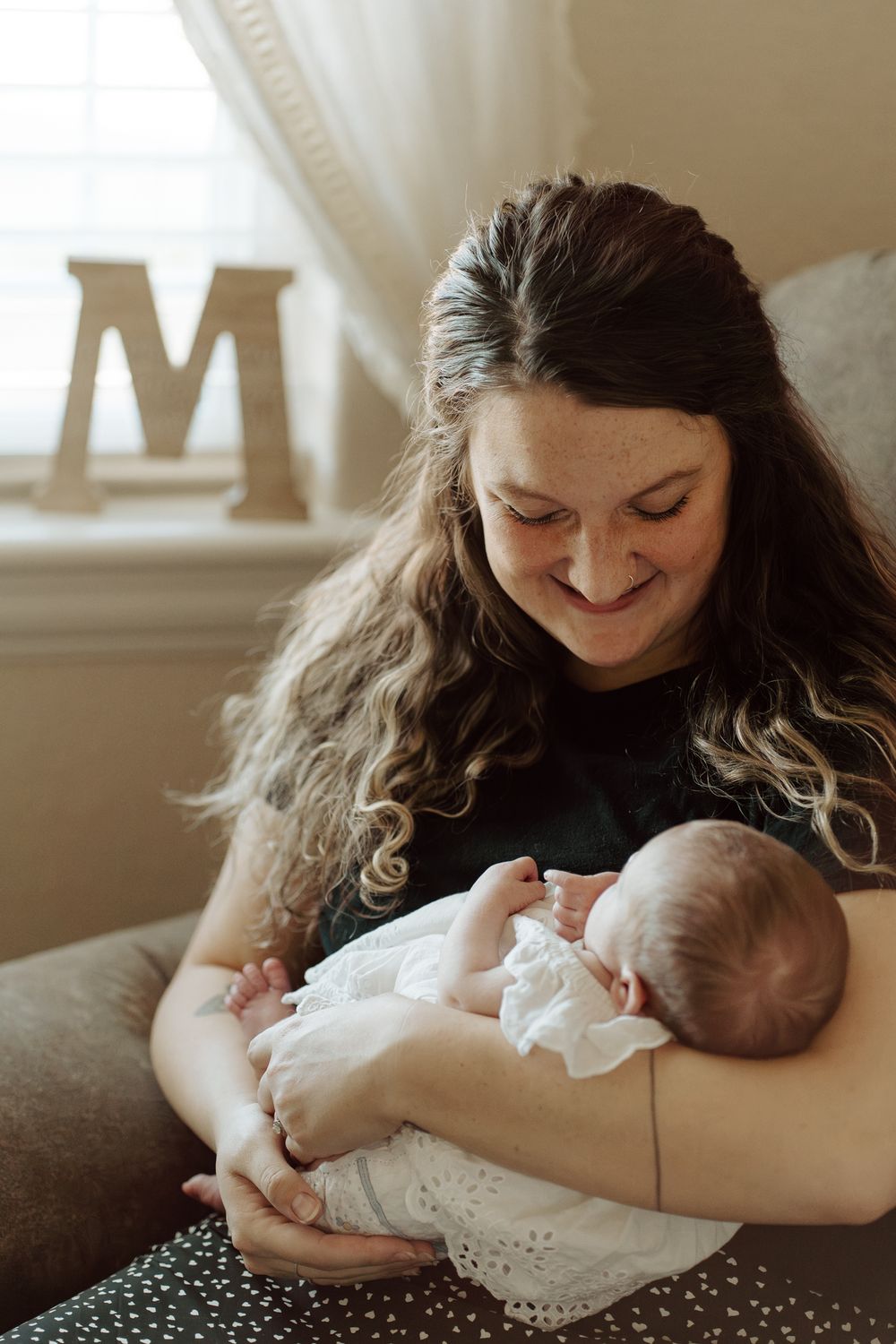Smiling mother with long curly hair cradling newborn baby in white dress, seated in nursery chair near decorative letter M.