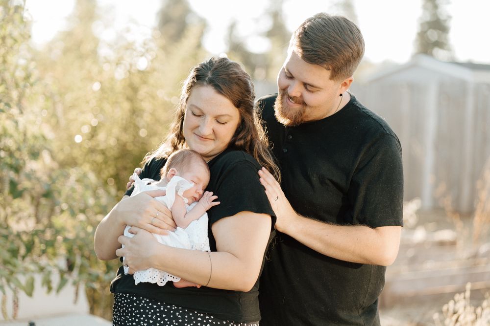 A young couple smiles while holding their newborn baby outdoors in warm golden light.