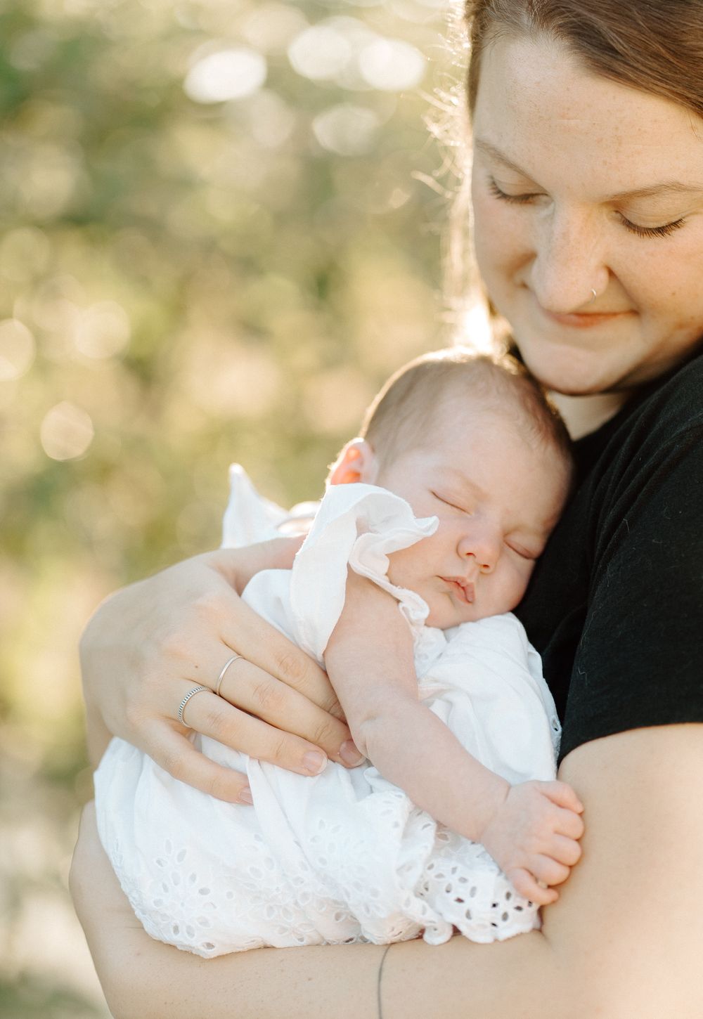A mother tenderly holds her sleeping newborn dressed in white lace outdoors.