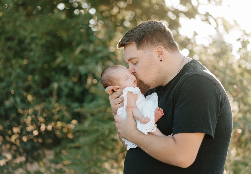 A father tenderly kisses his newborn baby outdoors, surrounded by lush green trees in warm sunlight.