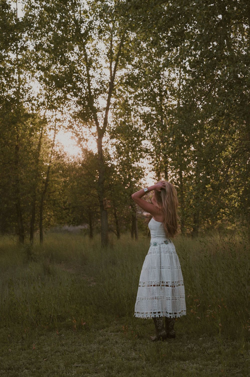 Woman in white dress and boots stands in a sunlit forest clearing at golden hour, hair blowing gently.