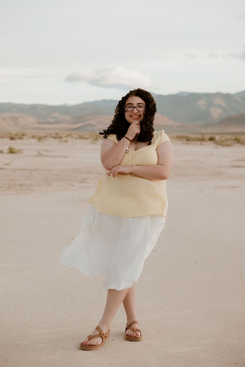 Woman in yellow top and white skirt posing in desert landscape with mountains in background.