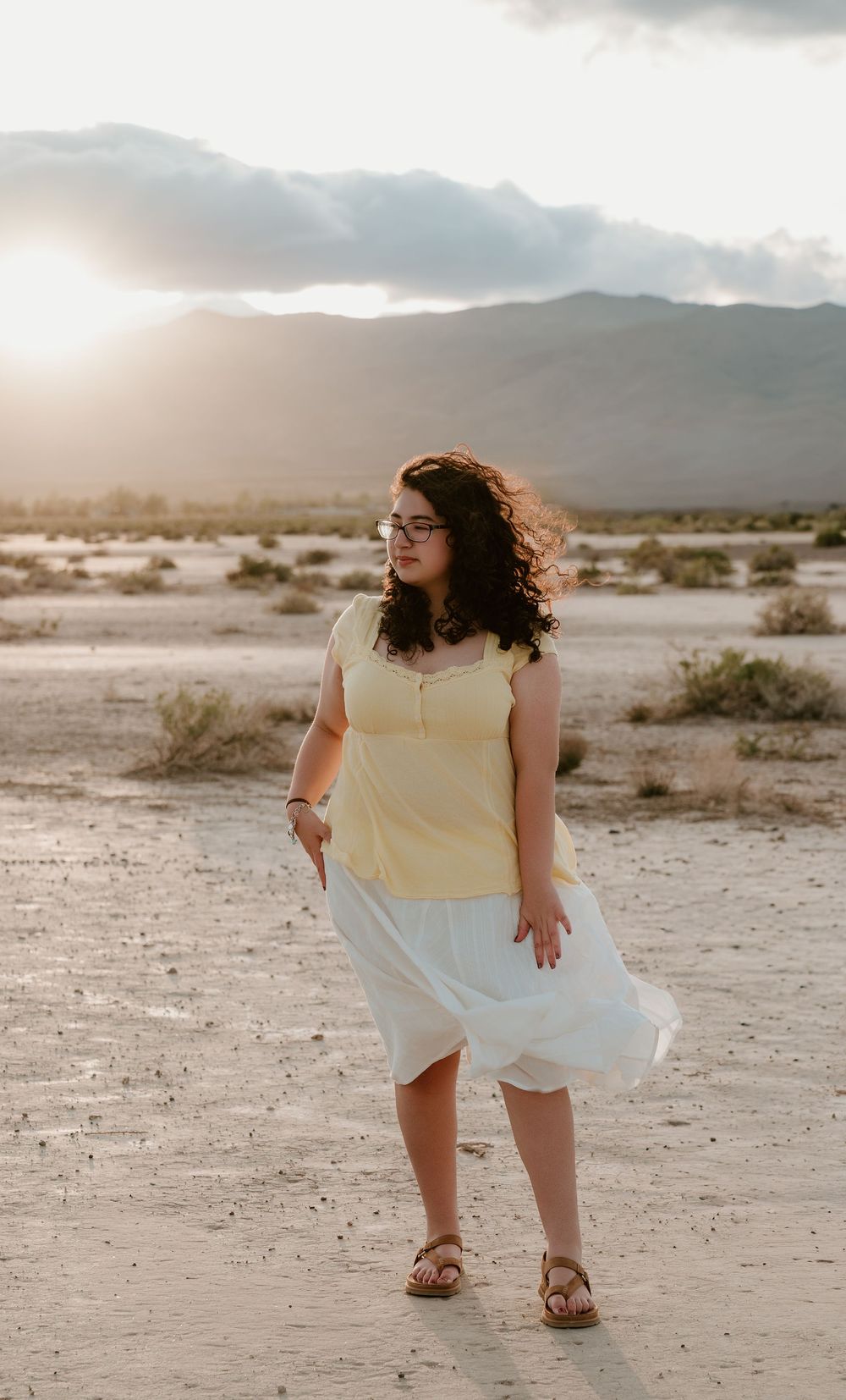 Woman in yellow top and white skirt stands in desert landscape at golden hour with mountains in background.