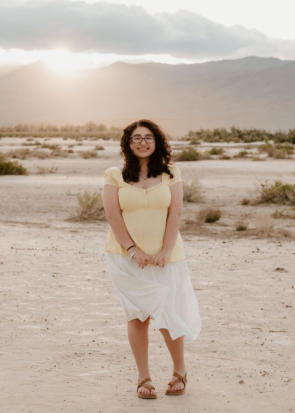 Woman in yellow top and white skirt poses in desert landscape at golden hour with mountains in background.