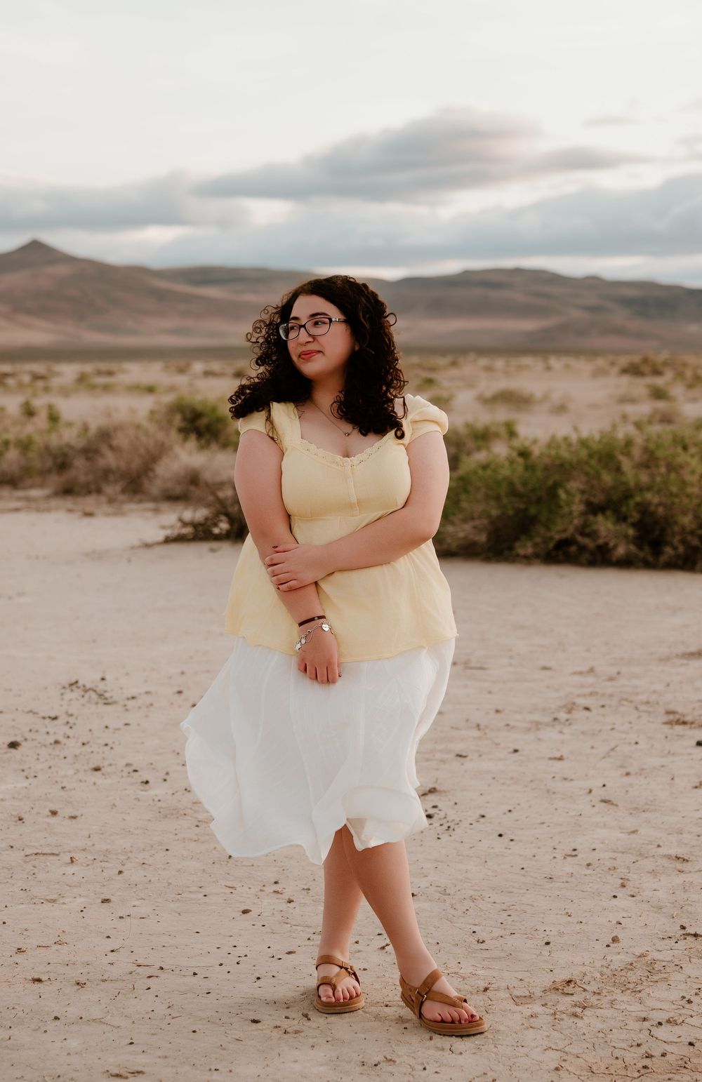 Young woman in yellow top and white skirt poses in desert landscape with mountains in background.