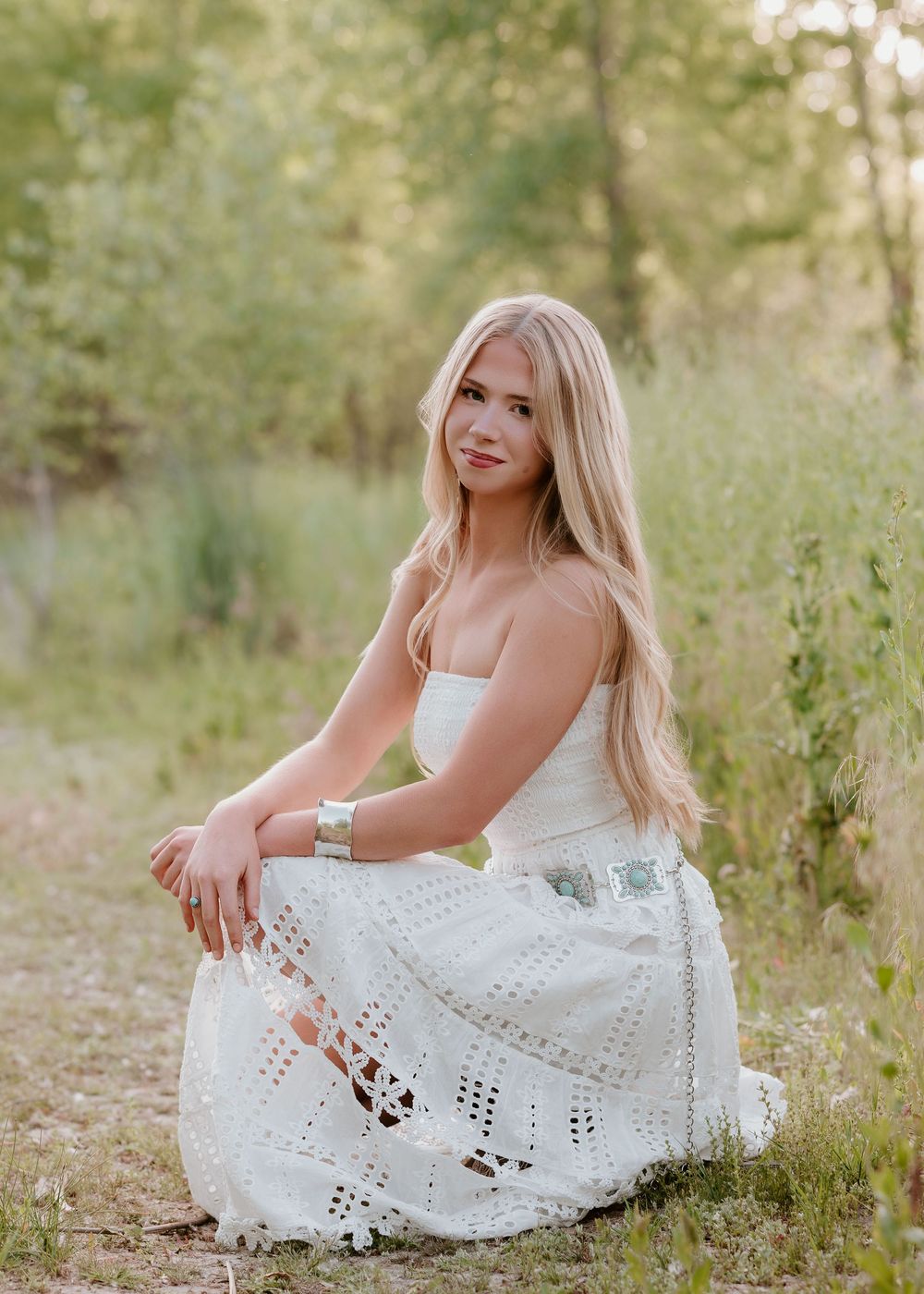 Young blonde woman in white eyelet dress sitting gracefully outdoors in a lush green natural setting.