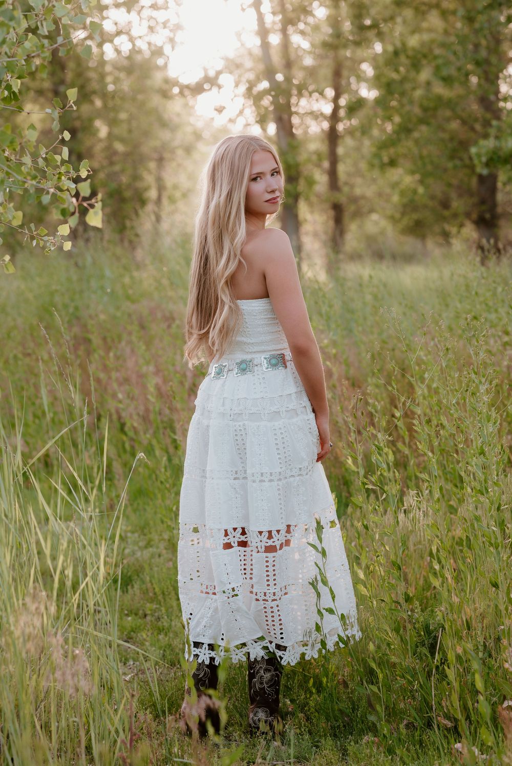 Young blonde woman in white lace midi dress and cowboy boots standing in a sunlit forest meadow.