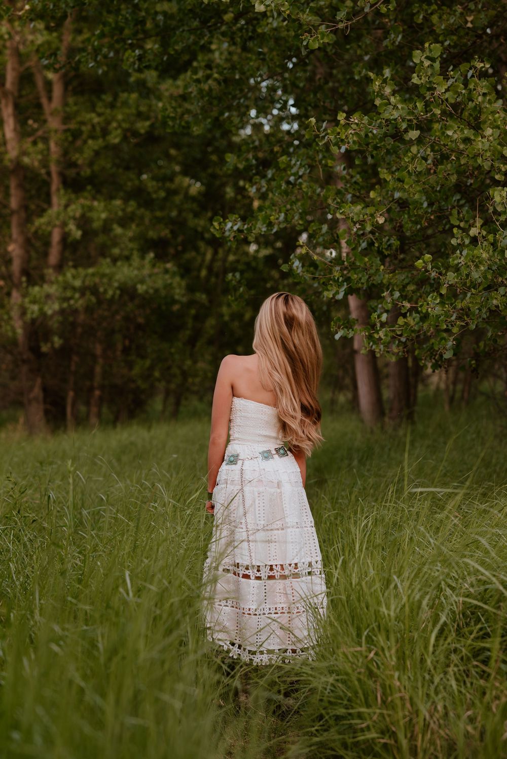 A woman in a white strapless lace maxi dress stands in tall green grass near lush trees.