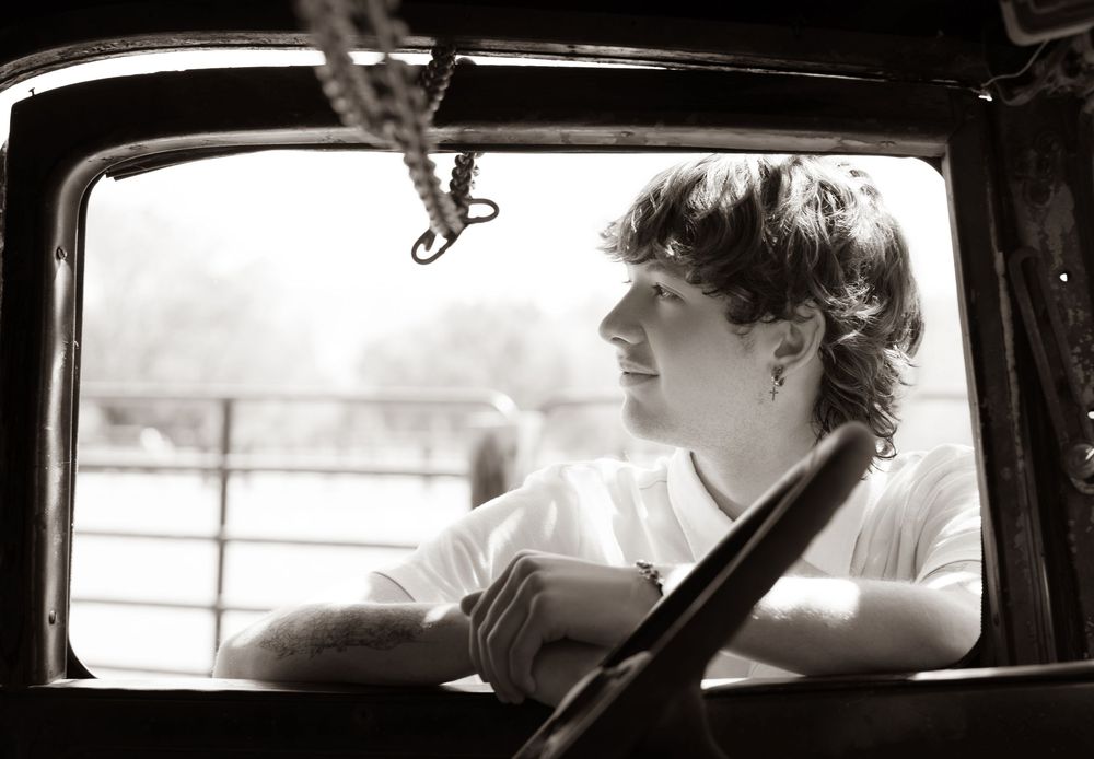 Black and white photo of a young man leaning on a vintage truck window, gazing thoughtfully into the distance.