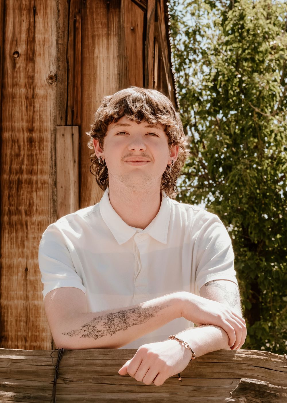 Young man with curly hair and tattoos leans on wooden fence, smiling in sunlight outdoors.