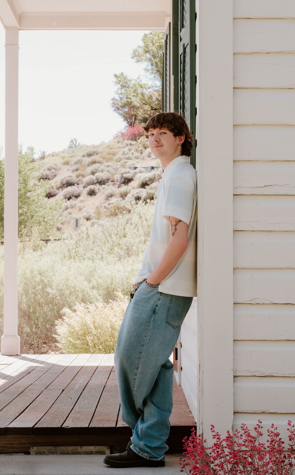 Young person in white t-shirt and baggy jeans leaning against white house exterior with scenic hillside backdrop.