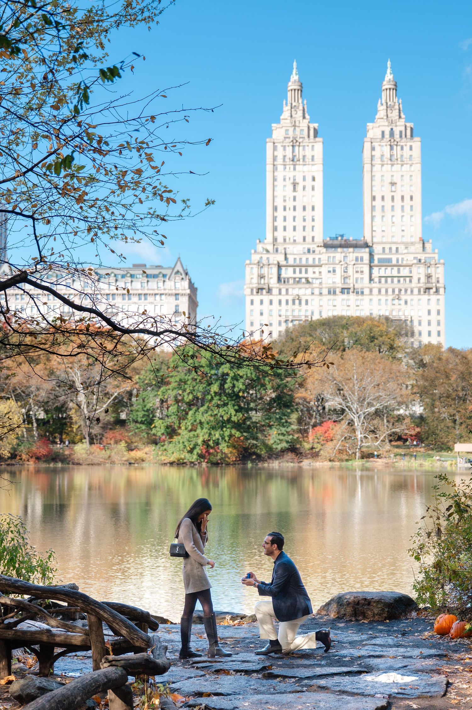 Man proposes to woman by Central Park lake with San Remo building towers in autumn backdrop, New York City.