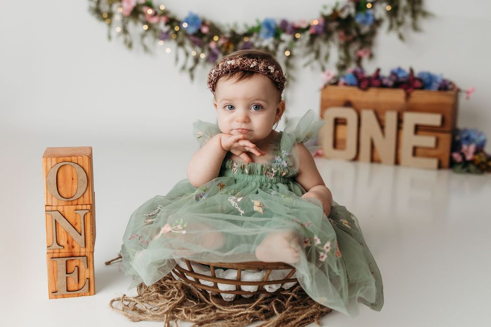 Baby girl in green dress sits in wicker basket surrounded by ONE signs for her first birthday cake smash photo.