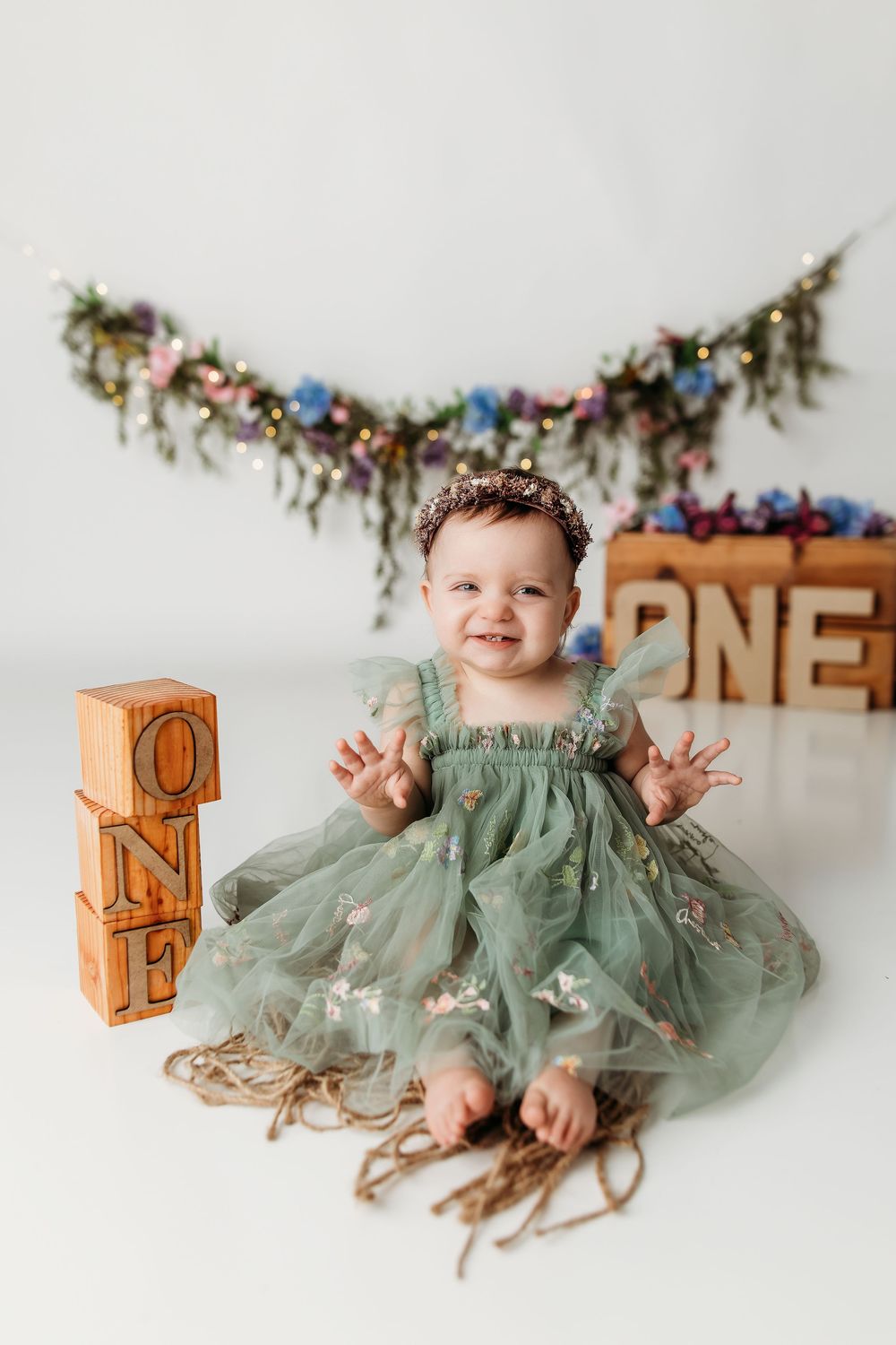 Baby girl in green tulle dress sits smiling near ONE blocks and floral garland during first birthday photo session.