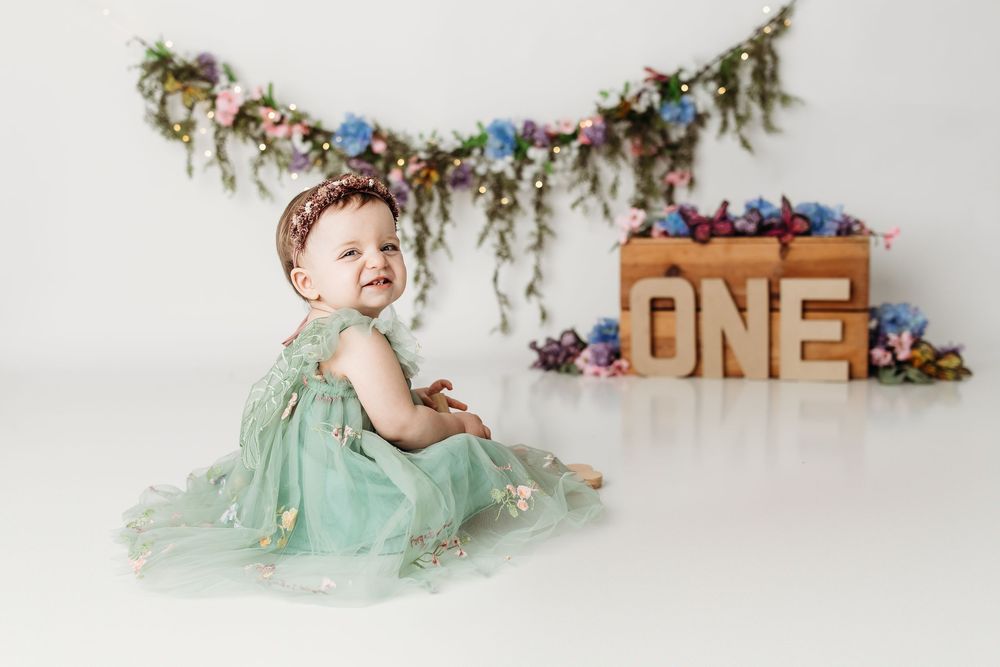 Smiling one-year-old baby girl in a green tulle dress sitting near a floral garland and wooden ONE sign.