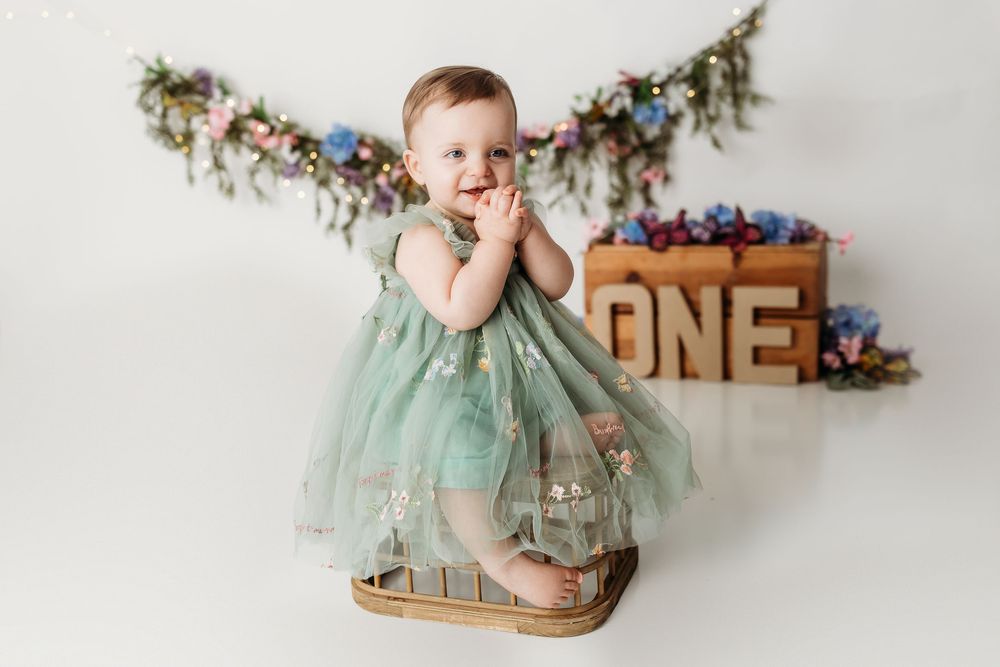Baby girl in green tulle dress stands in basket at ONE birthday cake smash photo session.