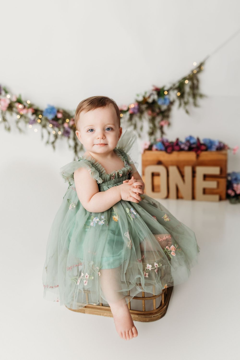 A baby girl in a green tulle dress sits in a basket celebrating her first birthday with floral decorations.