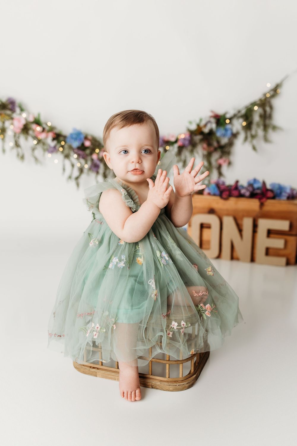 Adorable one-year-old baby girl in embroidered green tulle dress clapping hands in front of floral garland backdrop.
