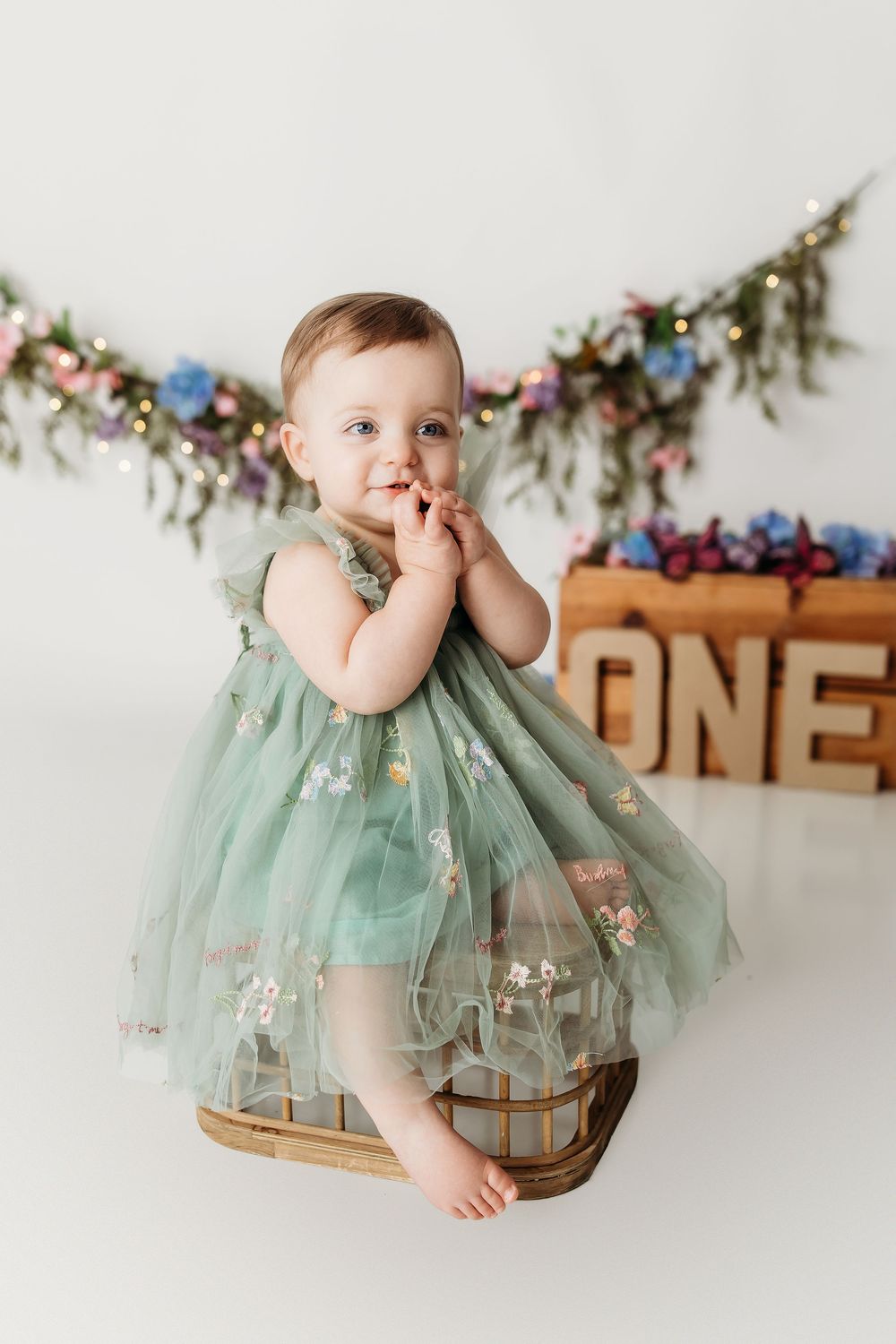 A baby girl in a green floral tulle dress sits in a basket, celebrating her first birthday with ONE sign decor.