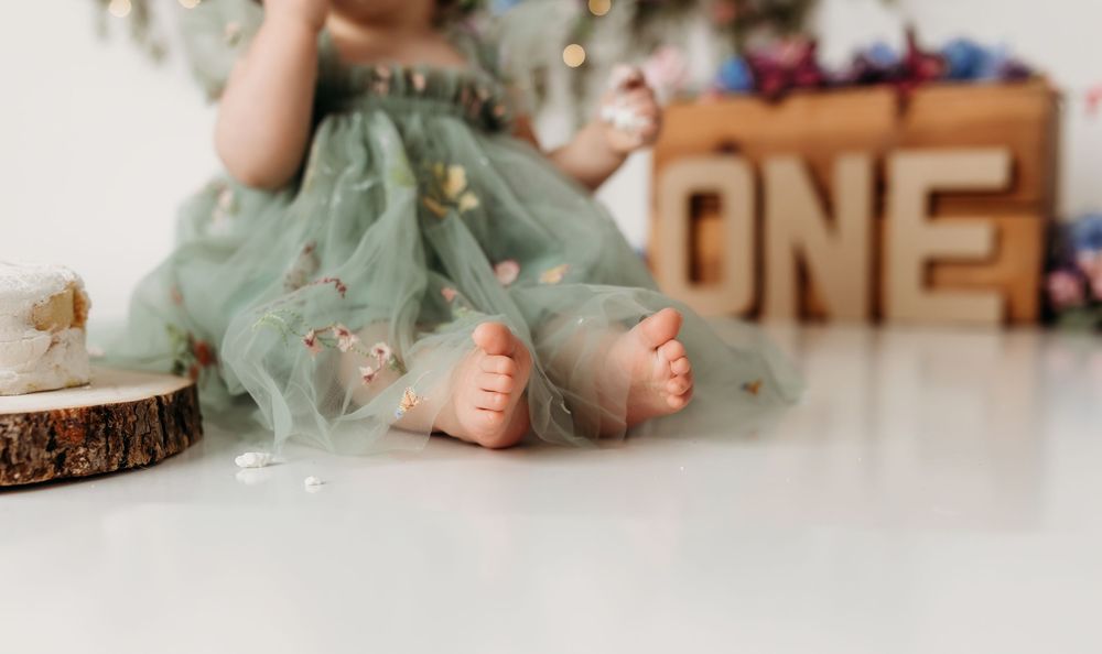 Baby girl in green tulle dress sits on white floor beside wooden ONE sign at first birthday cake smash photo session.