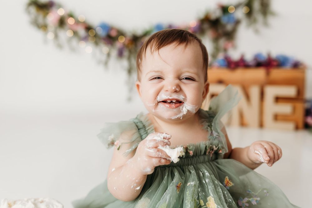 Smiling baby girl with cake on face enjoys first birthday cake smash in green floral dress.