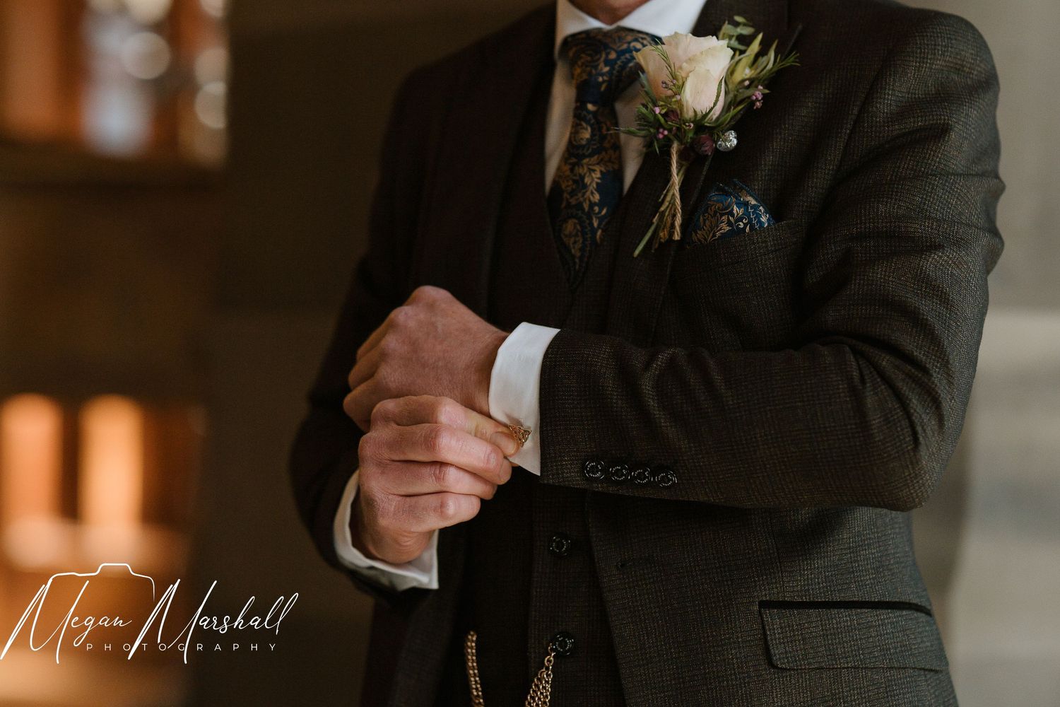 Groom in a dark suit adjusting cufflinks, wearing a floral boutonniere and pocket square at a wedding.