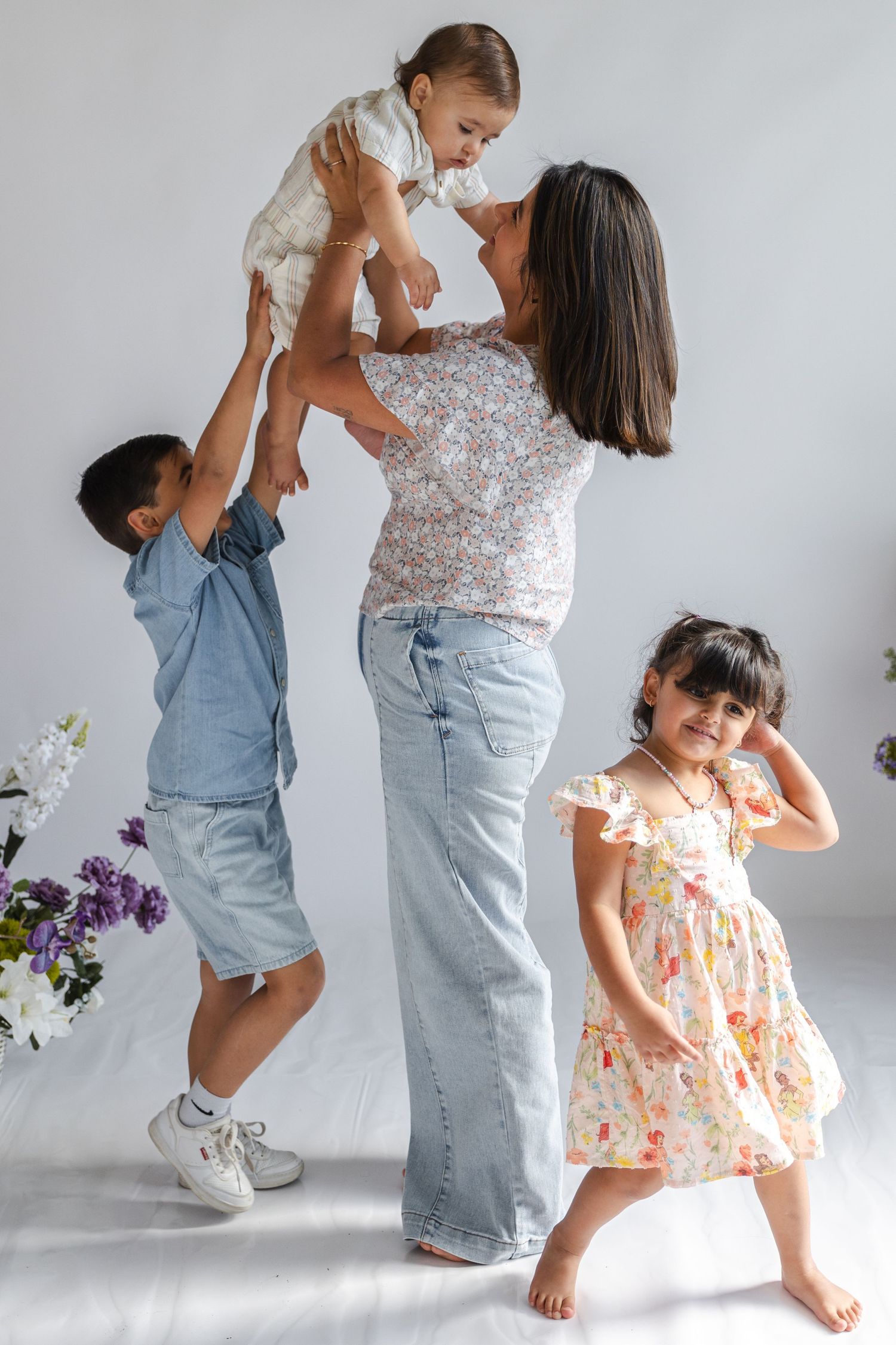 A joyful mother lifts a baby while two young children play around her in a bright, white studio setting.
